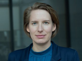 A close-up portrait of a person with short, light brown hair, wearing a blue blouse and a dark blazer, taken against a neutral background.