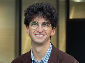 A young man with curly dark hair and glasses smiles at the camera, wearing a brown jacket and a striped shirt, against a blurred background.