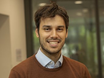 A smiling man with dark hair wearing a brown sweater over a blue shirt, standing indoors in front of a blurred background.