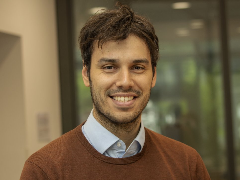 A smiling man with dark hair wearing a brown sweater over a blue shirt, standing indoors in front of a blurred background.