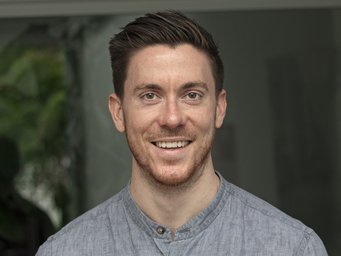A smiling man with short, dark hair and a light blue shirt, standing in a bright indoor setting.
