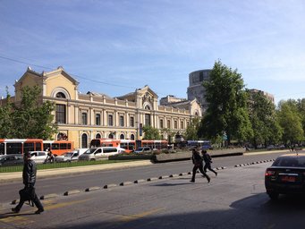 A street scene featuring a historic building on one side, city buses and cars, people crossing the street, and trees lining the road under a clear blue sky.