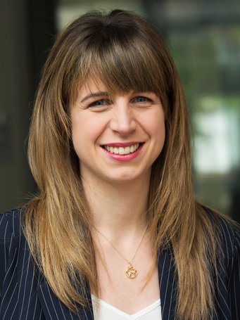 A person with long brown hair wearing an elegant dark blue pinstriped blazer and a gold necklace with a round pendant.