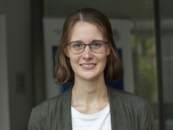 A woman with shoulder-length brown hair is wearing a gray jacket and a white T-shirt. She is standing in front of a modern office building with glass facades.