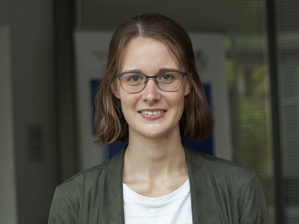 A woman with shoulder-length brown hair is wearing a gray jacket and a white T-shirt. She is standing in front of a modern office building with glass facades.