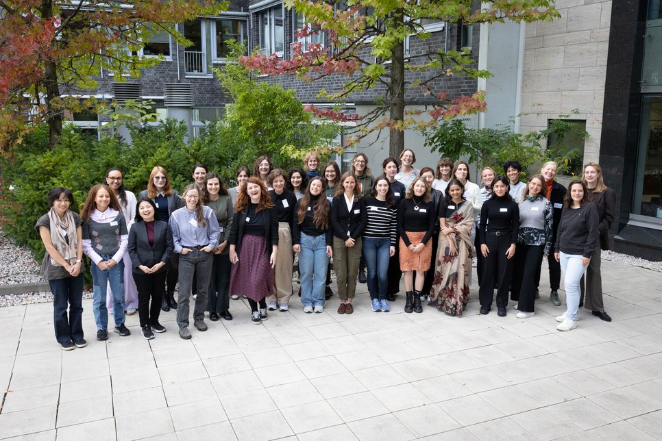 A diverse group of individuals is gathered outdoors, with greenery and trees in the background, standing on a paved area near a modern building facade.