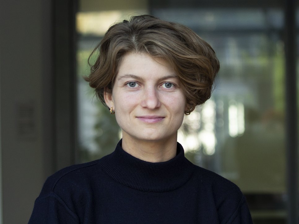 Woman with light brown short hair, wearing a dark sweater and gold earrings. Background slightly blurred with glass texture.