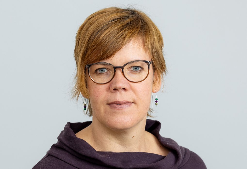 A woman with short brown hair, wearing glasses and a purple blouse, looking directly into the camera.