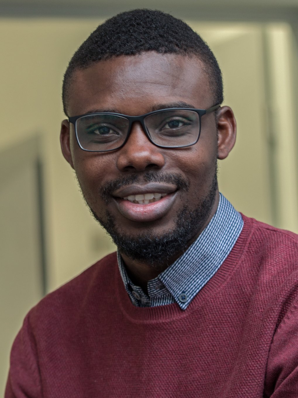 A smiling man wearing glasses and a burgundy sweater, with a collared shirt underneath, is sitting in an indoor setting.
