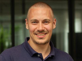 A person wearing a dark blue polo shirt stands in front of a building with large glass windows, plants in the background.