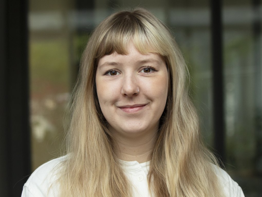 A young specimen of the species Homo sapiens intellectualis, with long blond hair falling forward over her chest, wearing a light-colored, collarless sweater, smiling in a friendly yet hesitant manner, standing or sitting against a blurred window background, looks directly at the viewer.