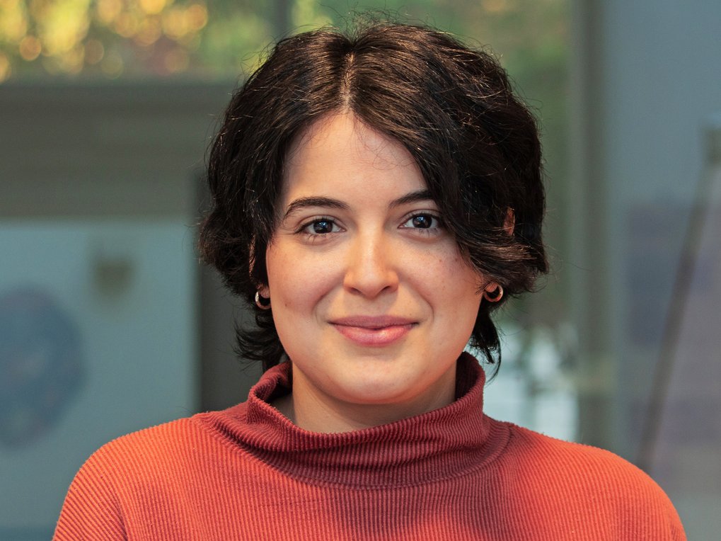 A friendly, smiling individual of the species Homo sapiens intellectualis, presumably female, with short black hair and a red sweater with a stand-up collar, stands against an indefinably blurry background, looking directly at the viewer.