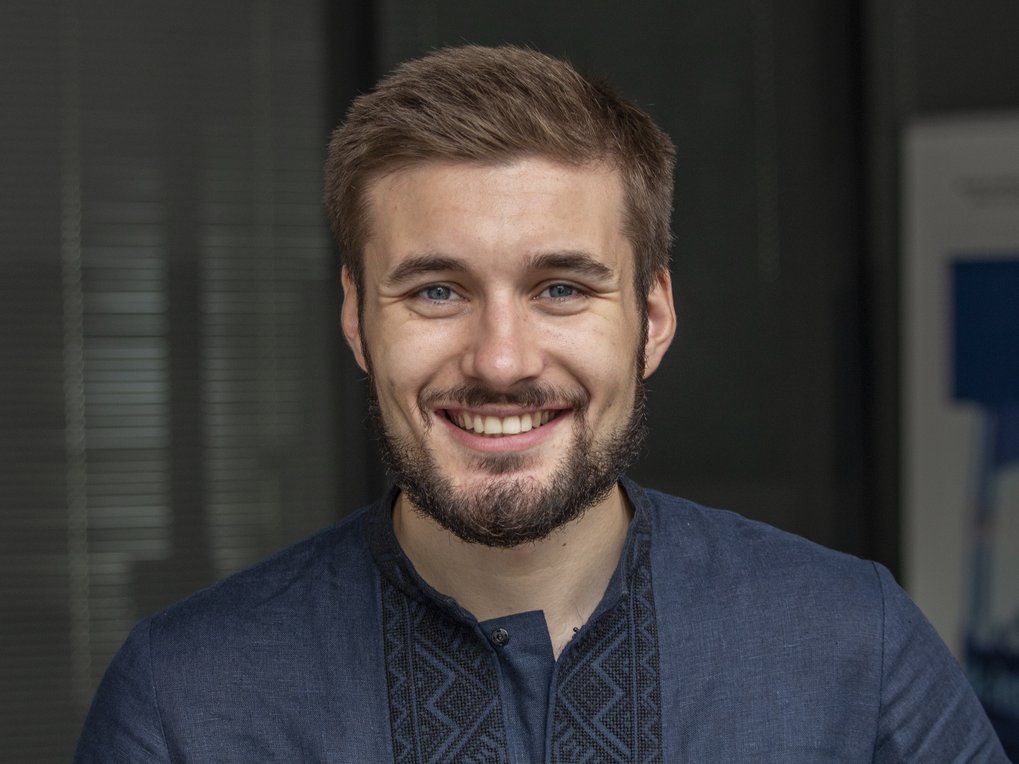 A friendly, smiling, bespectacled individual of the species Homo sapiens intellectualis, male, with thick, short hair and the hint of a budding beard, presents himself to the viewer. He is wearing an elegant dark blue collarless shirt and is standing or sitting in front of an indefinably blurry background.