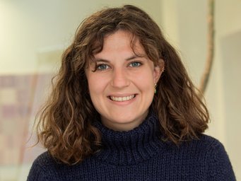 A woman with shoulder-length, curly brown hair is wearing a dark blue knit sweater and smiling directly at the camera; the background is out of focus.