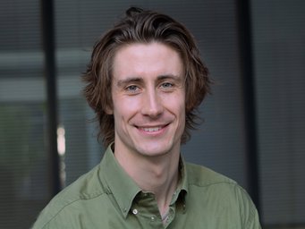 A young man with medium-length brown hair and a green shirt smiles at the camera; the background is out of focus and features vertical lines.