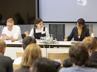 Work at the School pursues an empirical-analytical approach rather than an efficiency-theoretical, prescriptive one. Three panelists engaged in discussion at a conference, with audience members seated in the foreground.