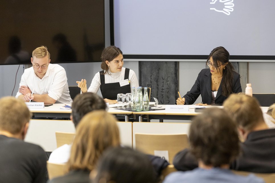 Three panelists engaged in discussion at a conference, with audience members seated in the foreground.