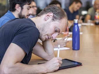 Students complete foundational coursework to strengthen their theoretical background and methodological skills. A person, focusing on writing on a tablet, is seated at a conference table.