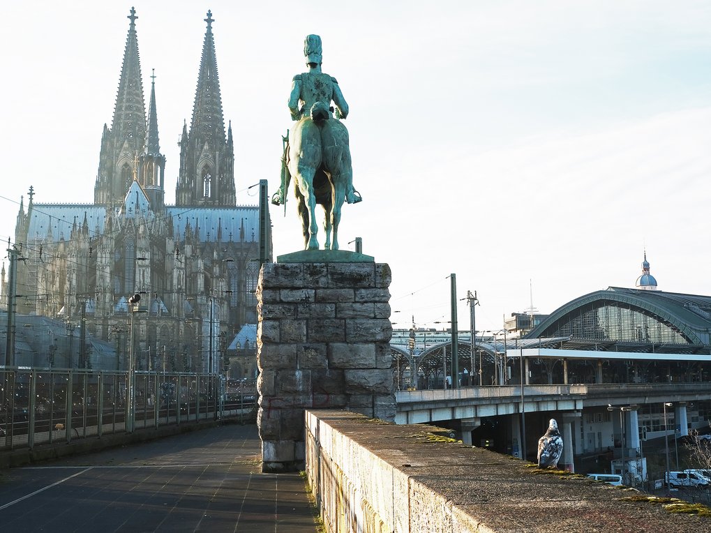 A statue of a horse rider on a pedestal, with a historic cathedral in the background, and a modern train station nearby.