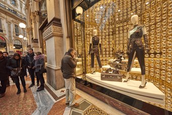 Eine geschäftige Einkaufspassage mit Menschen, die an einem Schaufenster eines luxuriösen Prada-Geschäfts vorbeigehen, in dem Schaufensterpuppen und Designerwaren ausgestellt sind.