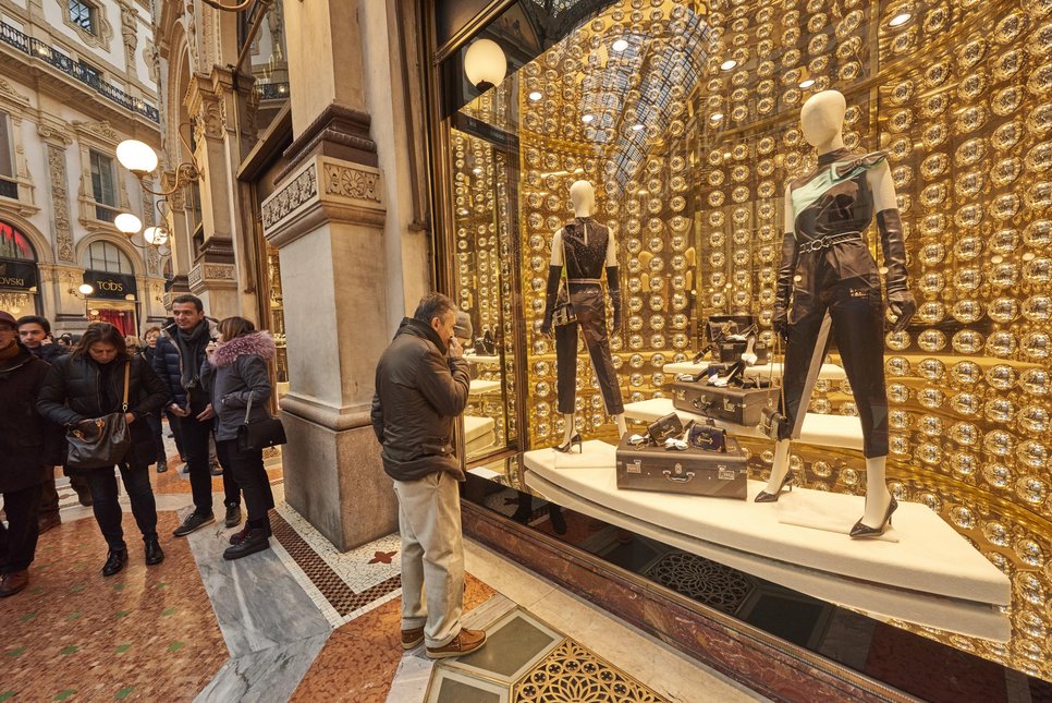 Eine geschäftige Einkaufspassage mit Menschen, die an einem Schaufenster eines luxuriösen Prada-Geschäfts vorbeigehen, in dem Schaufensterpuppen und Designerwaren ausgestellt sind.