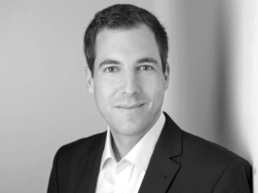 A professional portrait of a man in a suit, with a neutral expression, set against a plain background.