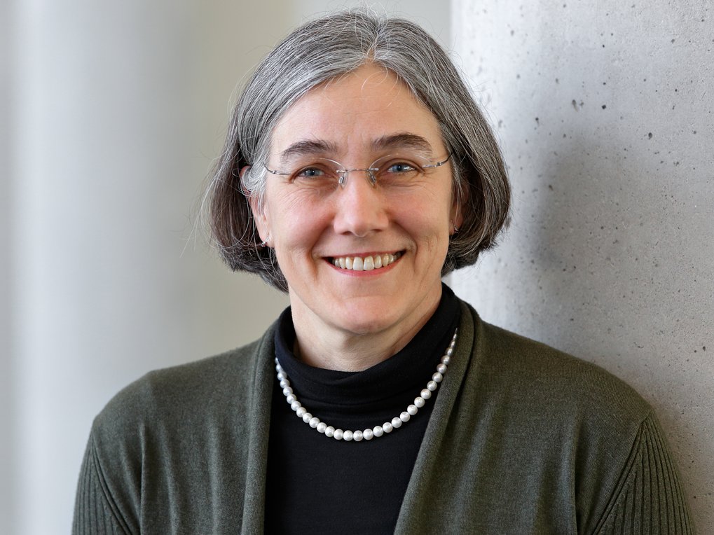 A woman with gray hair and glasses smiles while wearing a dark top and a pearl necklace, standing next to a concrete column.