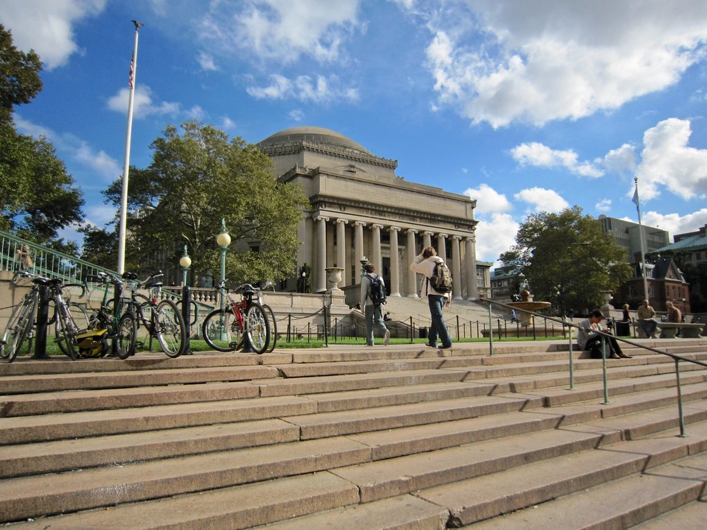 Universitätsgebäude im neoklassizistischen Stil mit Freitreppe davor, darauf ein paar Menschen und geparkte Fahrräder, blauer Himmel, weiße Wolken
