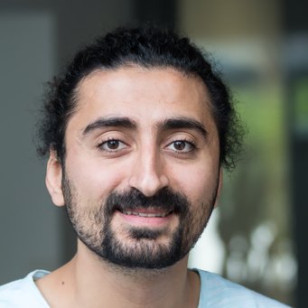 © MPIfG / Dünkelmann A smiling man with curly hair and a beard, wearing a light blue shirt, posed in an indoor setting.