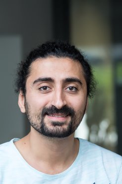 A smiling man with curly hair and a beard, wearing a light blue shirt, posed in an indoor setting.