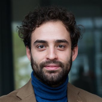 © MPIfG / Dünkelmann A man with curly hair and a beard, wearing a blue turtleneck and a brown blazer, smiling at the camera.