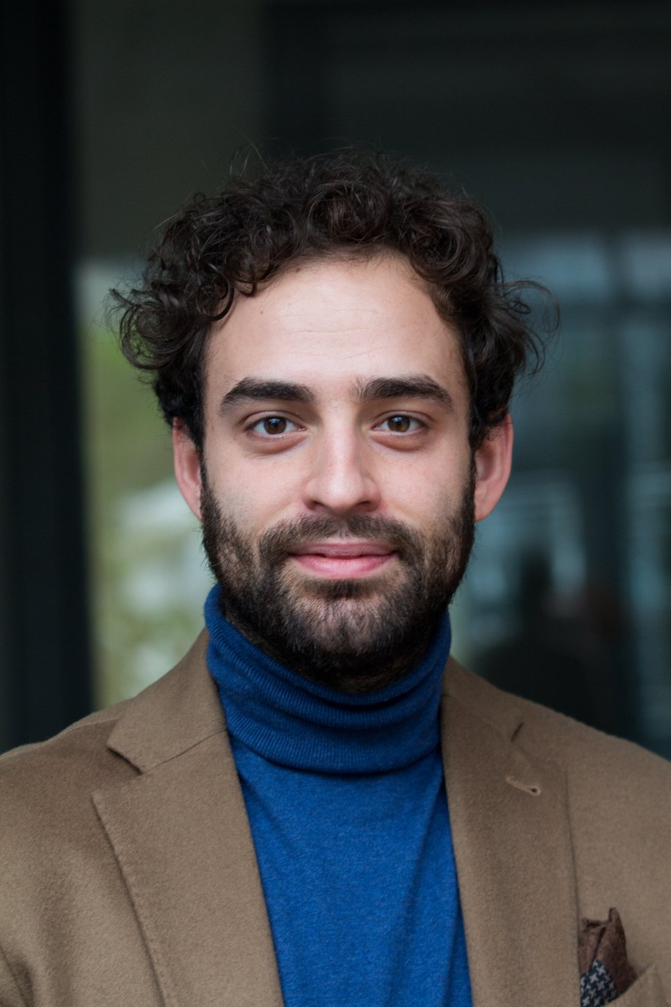 A man with curly hair and a beard, wearing a blue turtleneck and a brown blazer, smiling at the camera.