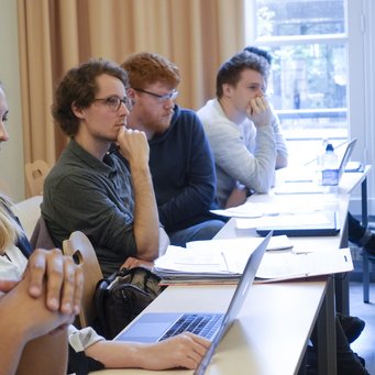 © MPIfG A group of students sitting at a table, engaged in a discussion or lecture, with laptops and notes in front of them.