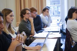 A group of students sitting at a table, engaged in a discussion or lecture, with laptops and notes in front of them.