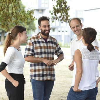 © MPIfG A group of four people engaged in a conversation outdoors, with trees and buildings in the background.