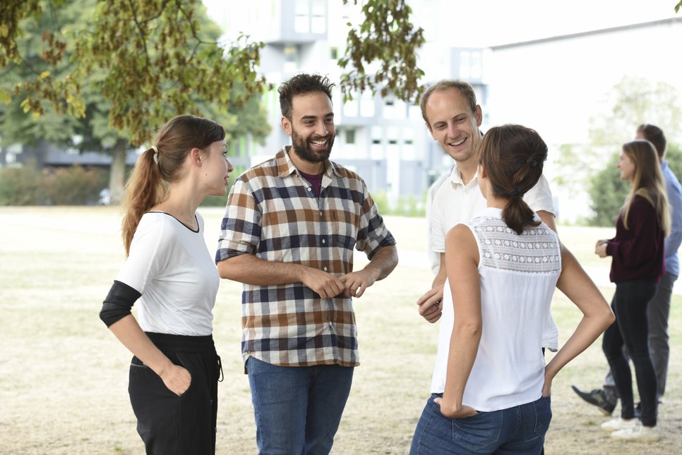 A group of four people engaged in a conversation outdoors, with trees and buildings in the background.