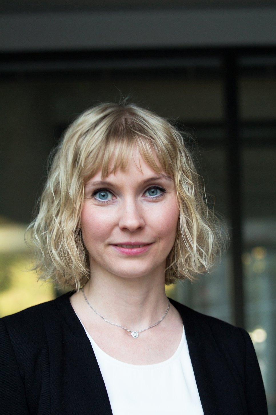 A woman with blonde, curly hair, wearing a black blazer, looks directly at the camera.