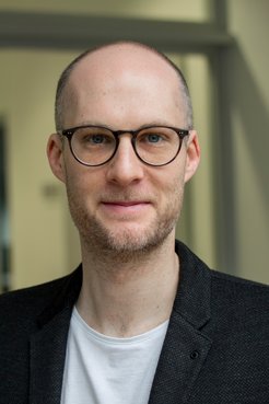 A man with a shaved head and glasses, wearing a dark jacket, smiles for the camera.