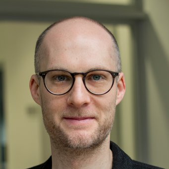 © MPIfG / Dünkelmann A man with a shaved head and glasses, wearing a dark jacket, smiles for the camera.