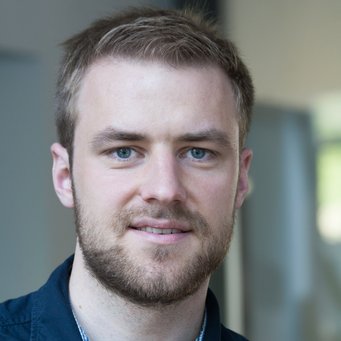 © MPIfG / Dünkelmann A young man with short light brown hair and a beard, wearing a dark jacket over a checked shirt, smiling at the camera.