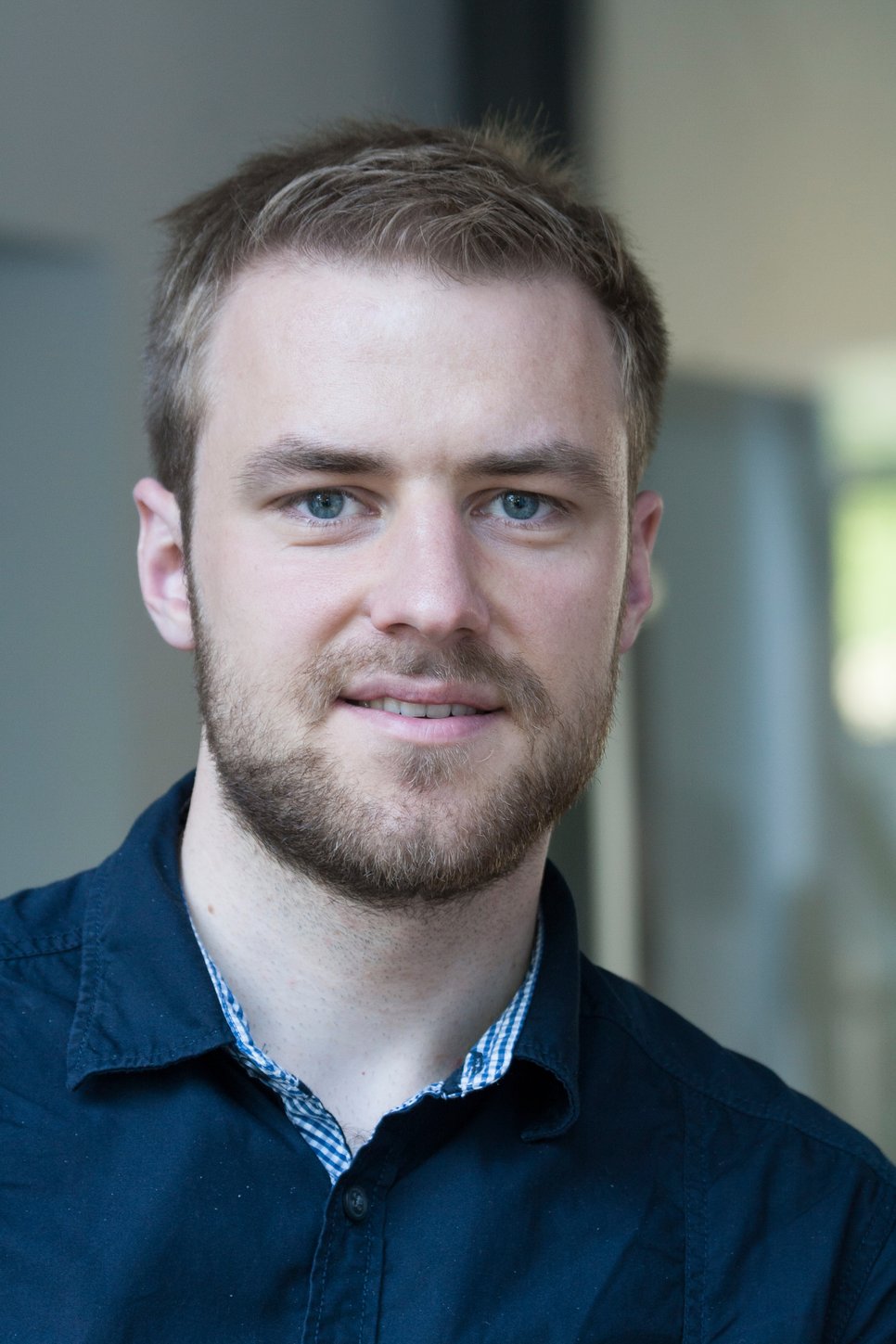 A young man with short light brown hair and a beard, wearing a dark jacket over a checked shirt, smiling at the camera.