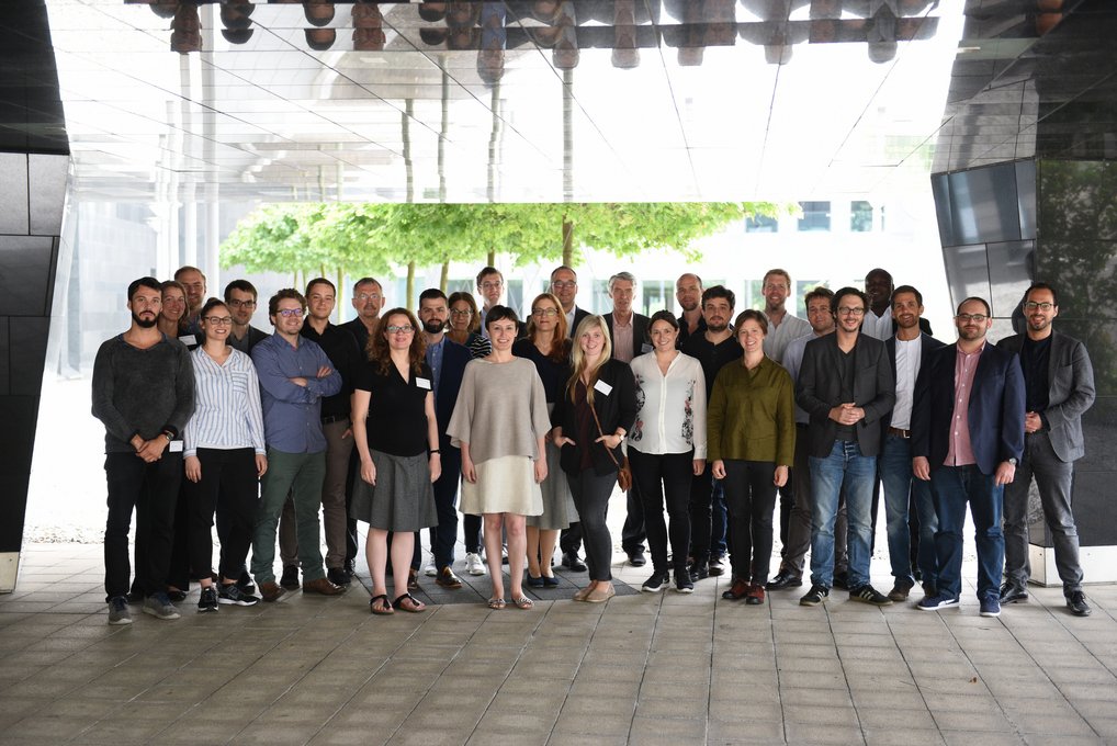 A group photo of approximately 30 people standing together under a modern architectural structure, with greenery visible in the background.