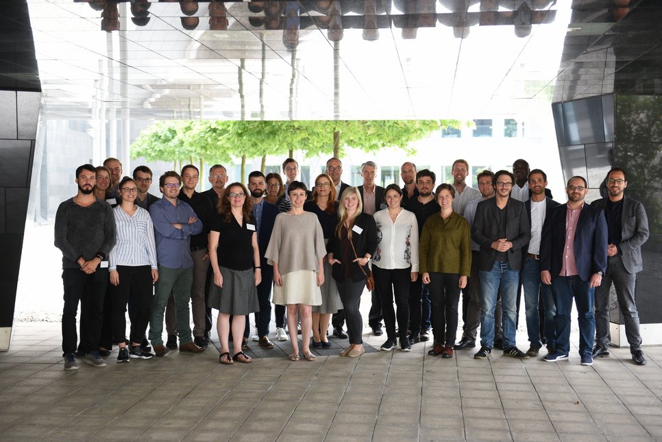 A group photo of approximately 30 people standing together under a modern architectural structure, with greenery visible in the background.