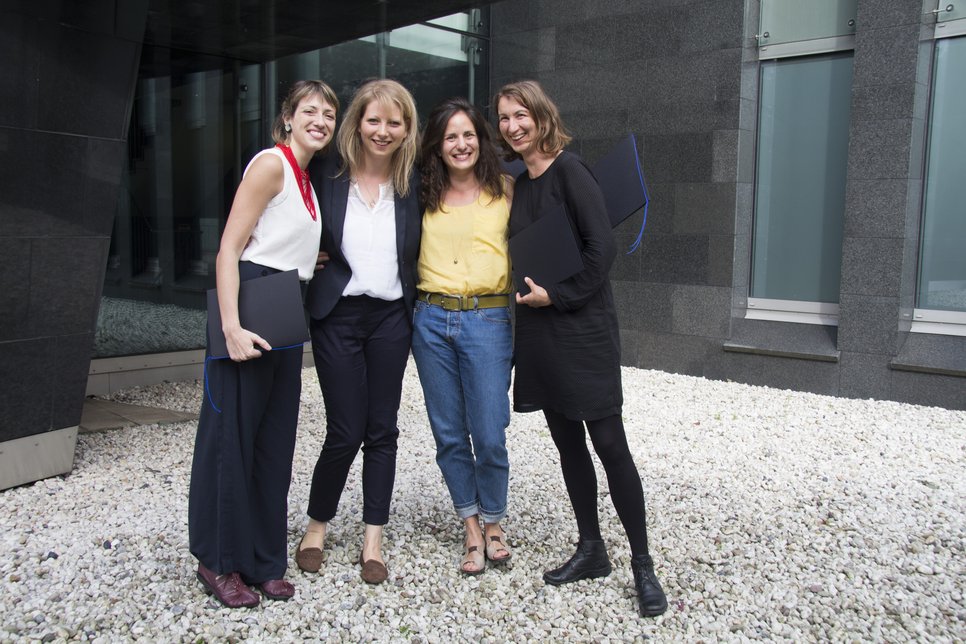 Four women standing together outdoors, smiling and holding black folders, against a backdrop of modern architecture and gravel.