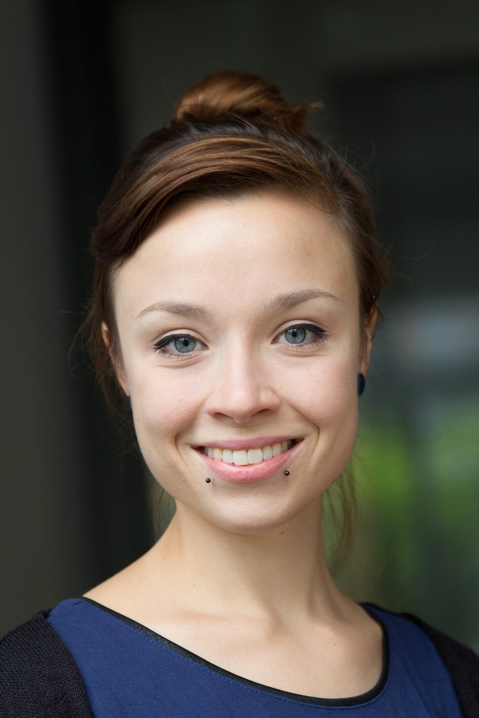 A close-up portrait of a woman with brown hair and blue eyes, smiling and wearing pierced earrings.