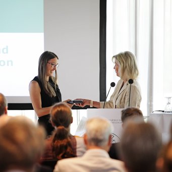 © MPIfG Two women are standing on stage, one handing an object to the other in front of an audience.