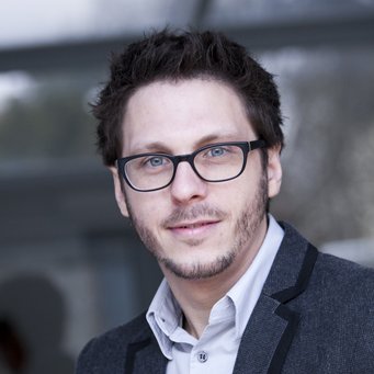 © MPIfG / Dünkelmann A man with short, dark hair and glasses, wearing a gray blazer over a light shirt, is smiling in a blurred outdoor setting.