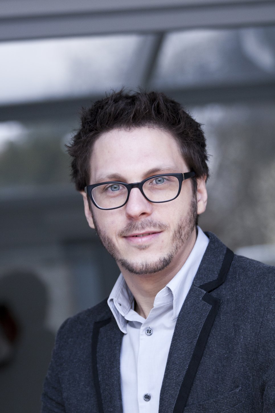 A man with short, dark hair and glasses, wearing a gray blazer over a light shirt, is smiling in a blurred outdoor setting.