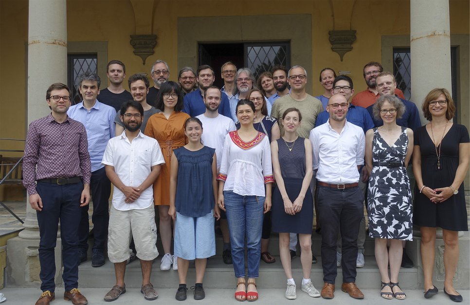 A group photo of approximately 30 people posing together outside a building, with some wearing casual attire and others in formal clothing.