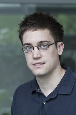 A young man with short dark hair and glasses, wearing a navy blue polo shirt, looking at the camera.